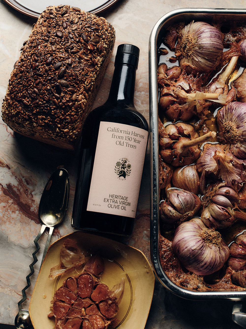 Artisanal whole grain bread next to a bottle of Heritage Extra Virgin Olive Oil, with a roasting pan filled with caramelized garlic cloves in the background.