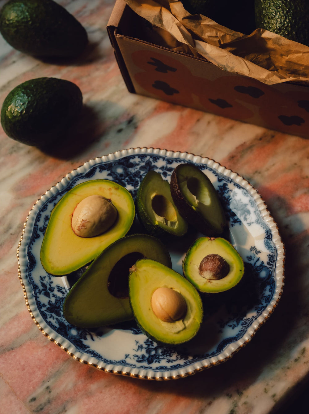 Halved avocados resting on a blue-patterned plate display their seeds, sitting on a marble surface.