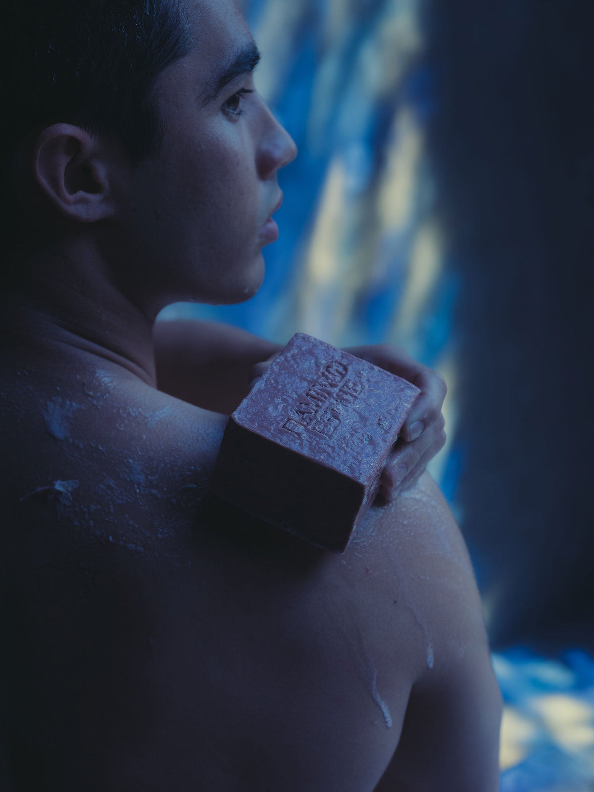 A person holds a brick-like bar of soap against their shoulder in a dim, blue-tinted room.