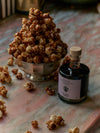 A metal bowl holds caramel popcorn beside a bottle labeled 