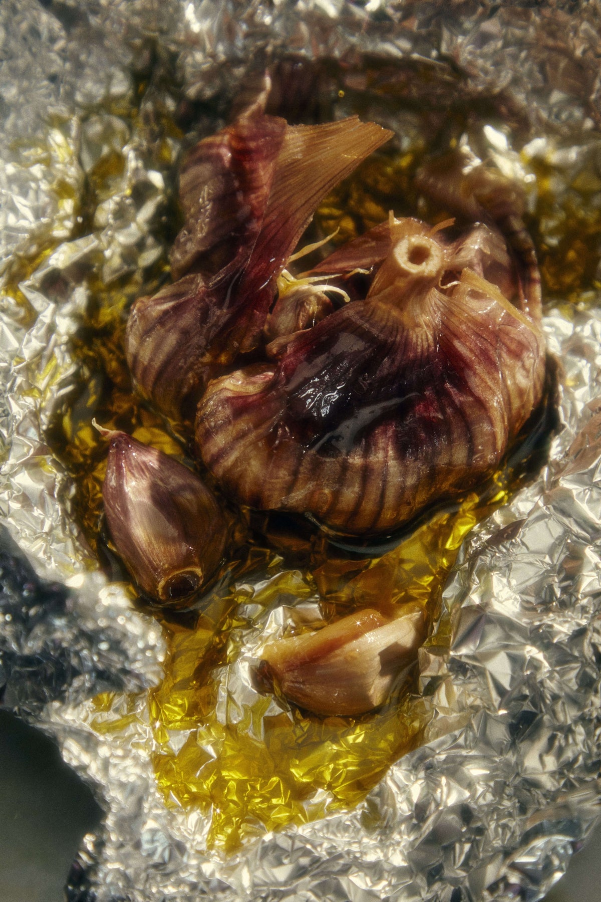 Close-up of roasted garlic cloves with skins, glistening in Heritage olive oil, wrapped in aluminum foil.
