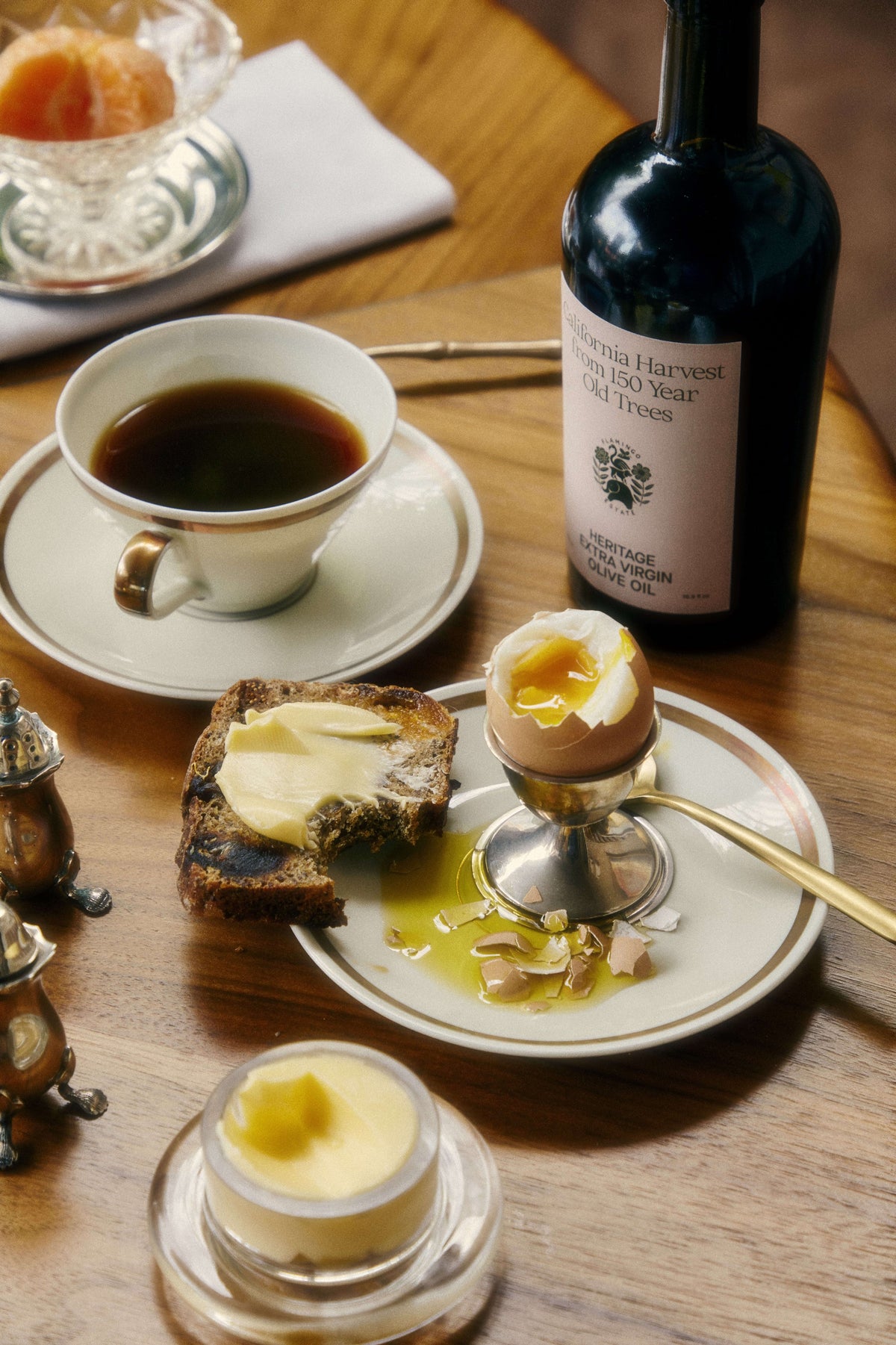A breakfast setting featuring a cup of black coffee, a slice of buttered toast, soft-boiled eggs, and a bottle of Heritage extra virgin olive oil.