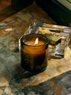A lit candle in a dark glass holder flickers on a marbled surface.