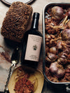 Artisanal whole grain bread next to a bottle of Heritage Extra Virgin Olive Oil, with a roasting pan filled with caramelized garlic cloves in the background.