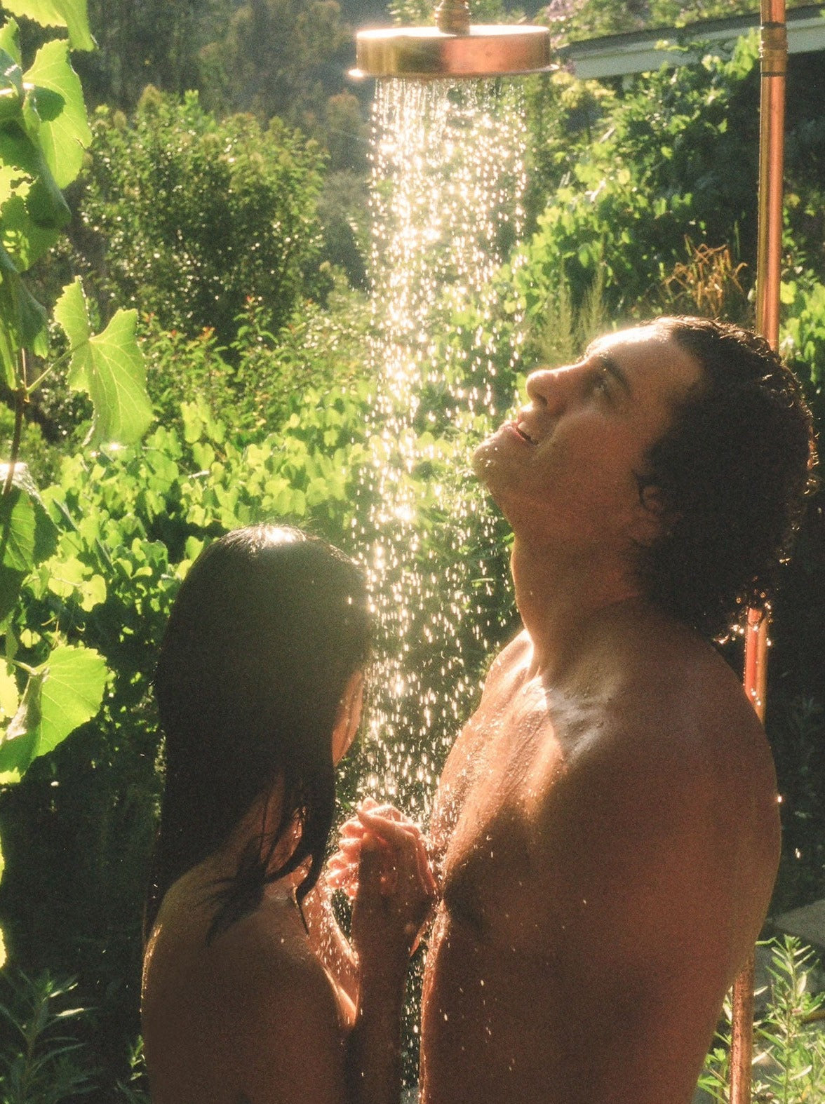 Man And Woman Showering Together Under An Outdoor Shower Surrounded By Greenery